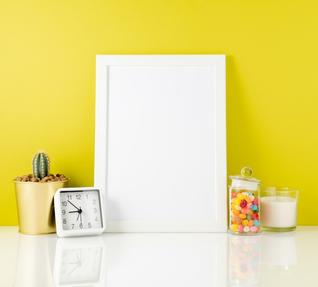 Blank white frame, clock, succulent, candy on a white table against the yellow wall