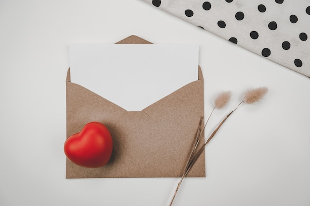 空白の白い紙は、赤いハートとウサギの尾のドライフラワーと白い布で開いた茶色の紙の封筒に置かれます