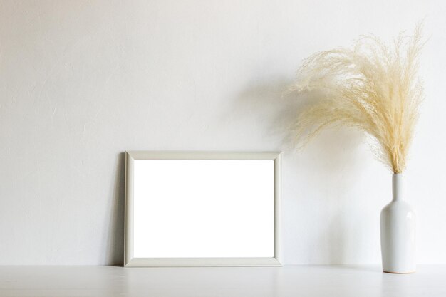 A blank white photo frame with brown natural flower on white vase over the white wall