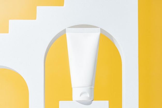 Blank white plastic cosmetics tube and white arch podium on\
bright yellow background skincare beauty treatment spa concept\
trendy showcase of cosmetics products