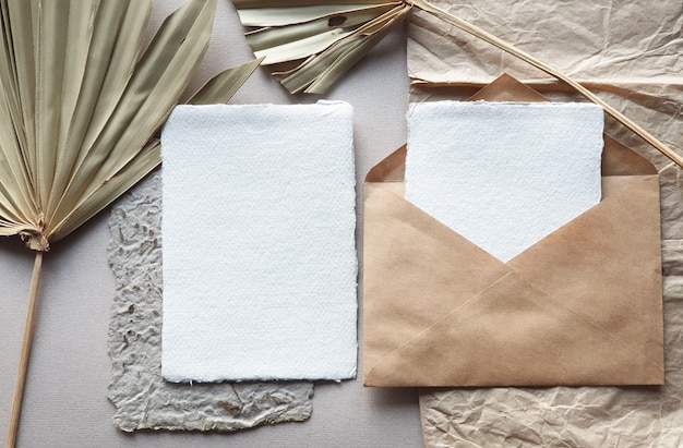 Blank white Wedding invitation cards mockups with dried palm leaf and craft envelope on textured table backgound. Elegant modern template for branding identity. Tropical design. Flat lay, top view