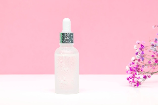 Blank witte glazen druppelflas verpakking met aromatische olie of serumwaterdruppels kleine bloemen twijg op roze achtergrondNatuurlijke biologische Spa Cosmetische productontwerp mockupVoorzijde vlakke laycopy ruimte