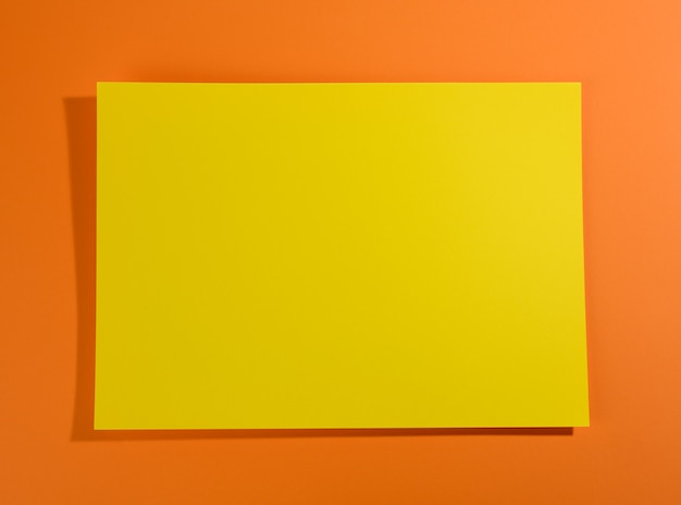 Blank yellow sheet of paper on orange surface with shadow, abstract backdrop for inscription, copy space