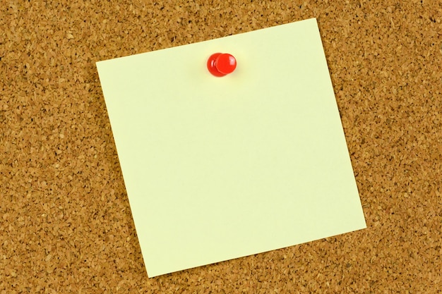 Blank yellow sticky note held on a cork notice board with a red pushpin