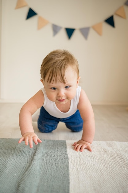 Foto blanke babyjongen in een witte bodysuit en spijkerbroek die op de vloer in de kinderkamer leert kruipen