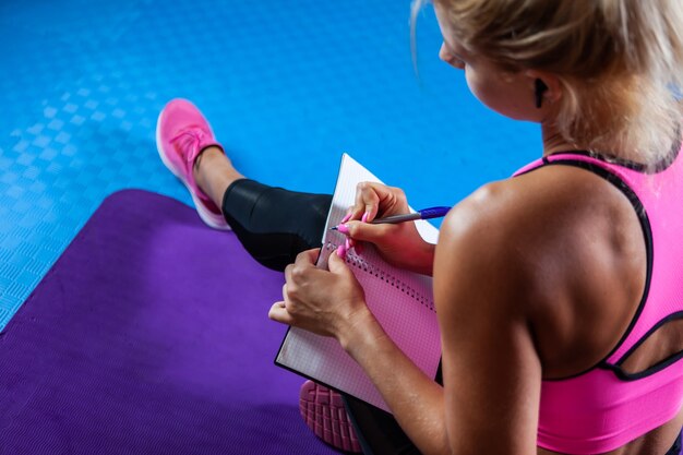 Blanke blonde fitnessvrouw denkt na en schrijft in een trainingsnotitieboekje een trainingsplan terwijl ze op een mat zit in een fitnessles. Achteraanzicht