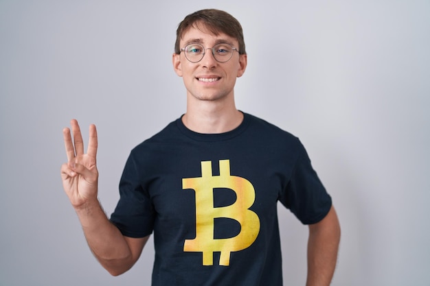 Blanke blonde man met bitcoin t-shirt die toont en omhoog wijst met vingers nummer drie terwijl hij zelfverzekerd en gelukkig glimlacht.