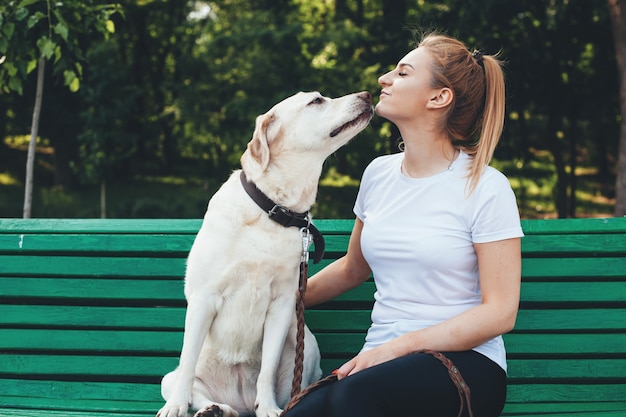 Blanke blonde vrouw en haar labrador zitten dicht op de bank en kussen tijdens een zomerse wandeling in het park