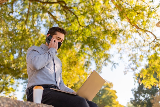 Blanke jonge man met een gezichtsmasker werken en studeren in engineering praten aan de telefoon en met behulp van zijn laptop in een prachtig park op een zonnige dag.