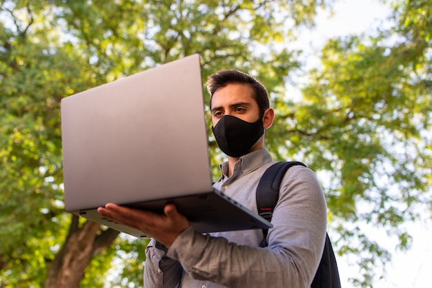 Blanke jonge man met gezichtsmasker werken en studeren in engineering met zijn laptop in een prachtig park op een zonnige dag