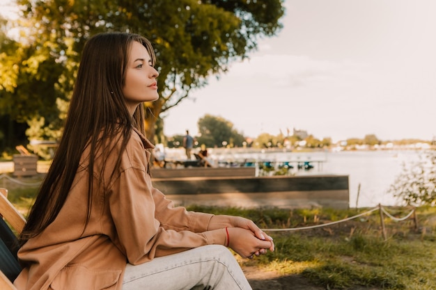 Blanke jonge mooie stijlvolle vrouw zittend op strandstoel pier in de buurt van rivier geklede bruine loopgraaf