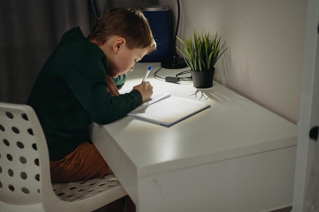 Blanke jongen doet huiswerk thuis in de donkere kamer bij het licht van de lamp. Afbeelding met selectieve focus