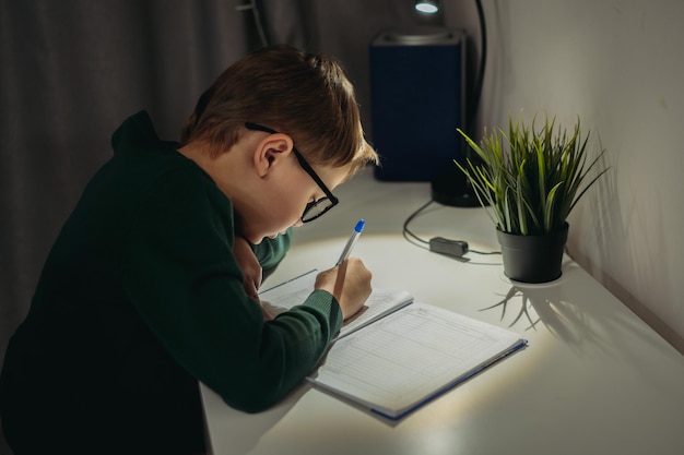 Blanke jongen doet huiswerk thuis in de donkere kamer bij het licht van de lamp