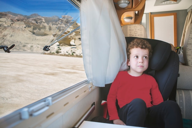 Foto blanke jongen met krullend haar zit bij het open raam van zijn camper met een spectaculair uitzicht