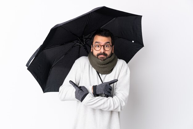 Blanke knappe man met baard met een paraplu over geïsoleerde witte muur wijzend op de zijkanten met twijfels