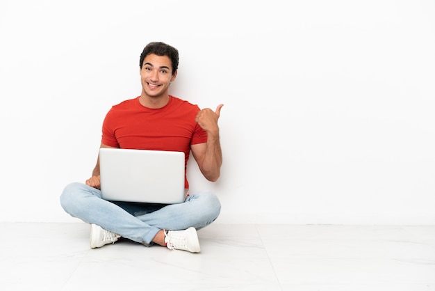 Blanke knappe man met een laptop die op de grond zit en naar de zijkant wijst om een product te presenteren