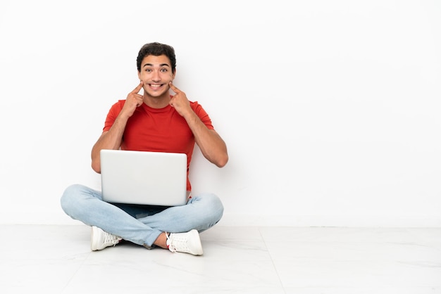 Blanke knappe man met een laptop zittend op de vloer glimlachend met een vrolijke en aangename uitdrukking