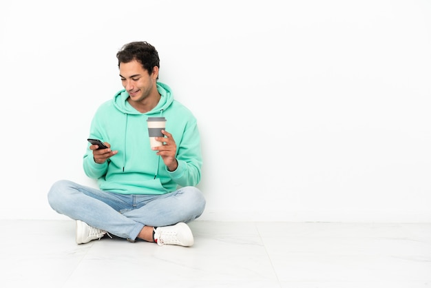 Blanke knappe man zittend op de vloer met koffie om mee te nemen en een mobiel