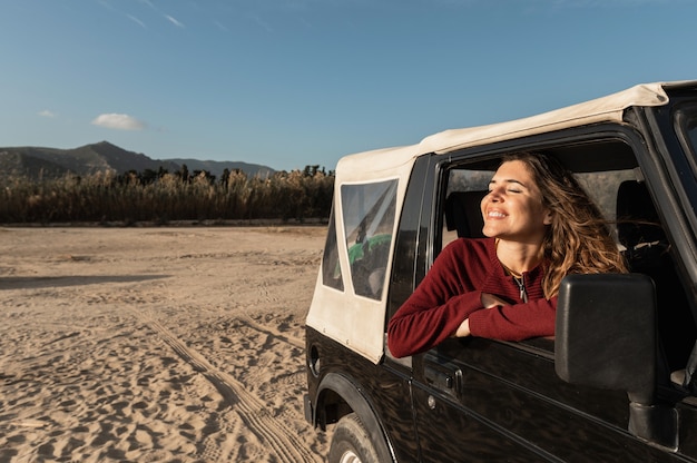 Blanke lachende jonge vrouw met gesloten ogen kijkt uit het raam. Reis in een off-road 4x4-voertuig. Achtergrond van zandwoestijn en strand.