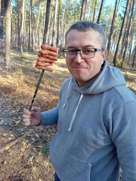 Blanke man eet hete gegrilde worst van een spies in het bos op een picknick