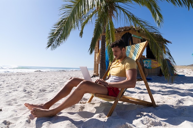 Blanke man geniet van tijd op het strand, zittend op een ligstoel, met behulp van een laptop