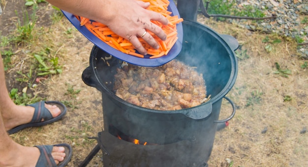 Blanke man kookt vlees met wortelen in een wokpan op een open vuur, zomer, buitenshuis