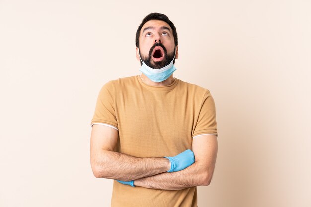 Blanke man met baard beschermen met een masker en handschoenen over muur opzoeken en met verbaasde uitdrukking