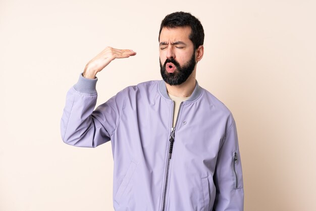 Blanke man met baard die een jas draagt over geïsoleerde muur met een vermoeide en zieke uitdrukking