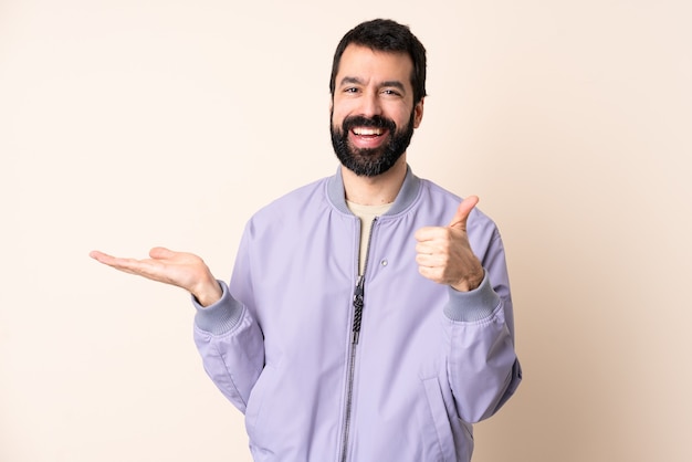 Blanke man met baard die een jas over geïsoleerde muur draagt die copyspace denkbeeldig op de palm houdt om een advertentie in te voegen en met omhoog duimen