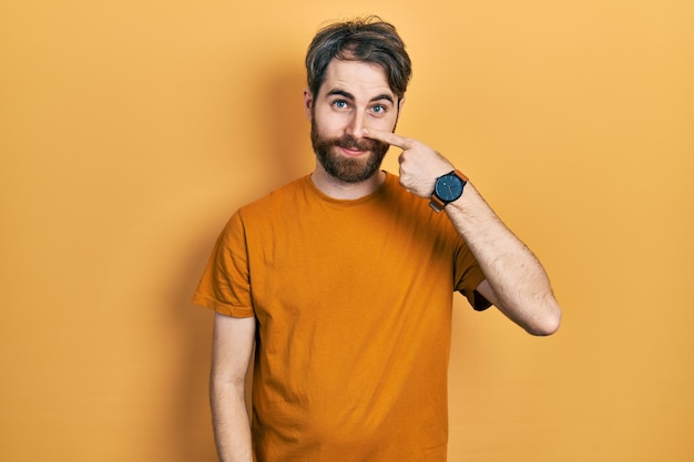 Blanke man met baard, gekleed in casual geel t-shirt wijzend met handvinger naar gezicht en neus lachend vrolijk schoonheidsconcept