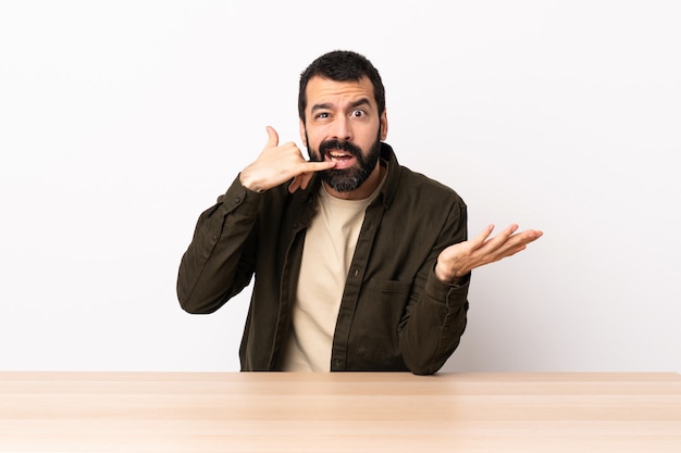 Blanke man met baard in een tafel telefoon gebaar maken en twijfelen