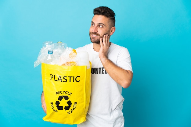 Blanke man met een zak vol plastic flessen om te recyclen geïsoleerd op blauwe achtergrond een idee denken tijdens het opzoeken