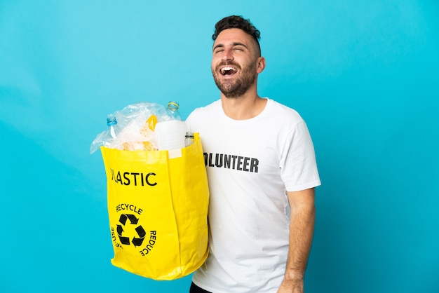 Blanke man met een zak vol plastic flessen om te recyclen geïsoleerd op blauwe achtergrond lachen