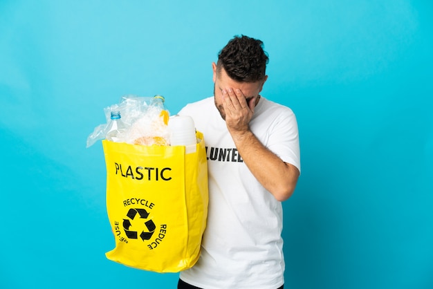 Blanke man met een zak vol plastic flessen om te recyclen geïsoleerd op blauwe achtergrond met vermoeide en zieke uitdrukking