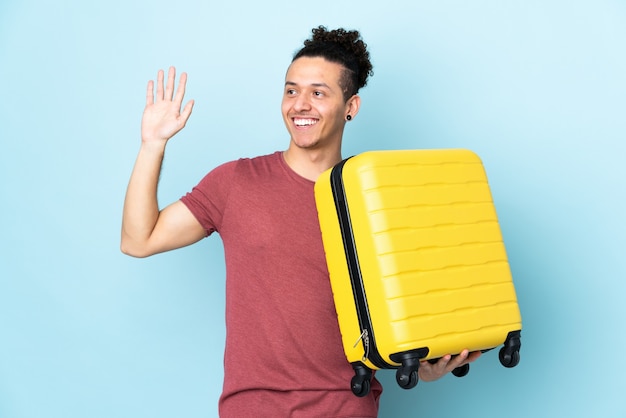 Blanke man over geïsoleerde blauwe achtergrond in vakantie met reiskoffer en groeten