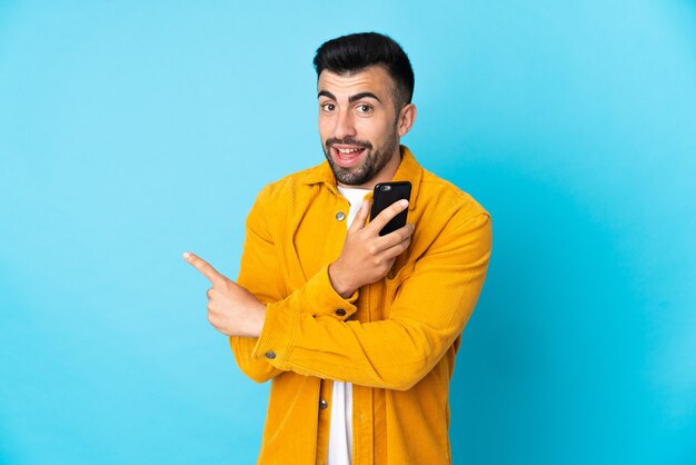 Blanke man over geïsoleerde blauwe muur met behulp van mobiele telefoon en terug te wijzen