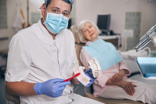 Blanke man-tandarts die op een kaakmodel laat zien hoe je de tanden goed schoonmaakt met een tandenborstel en een vrouw die naar de dokter luistert