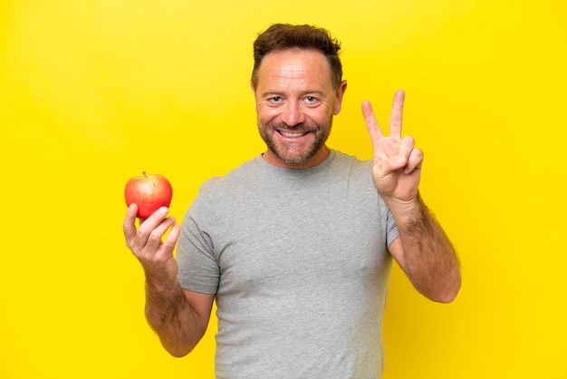 Blanke man van middelbare leeftijd met een appel geïsoleerd op gele achtergrond glimlachend en overwinningsteken tonend