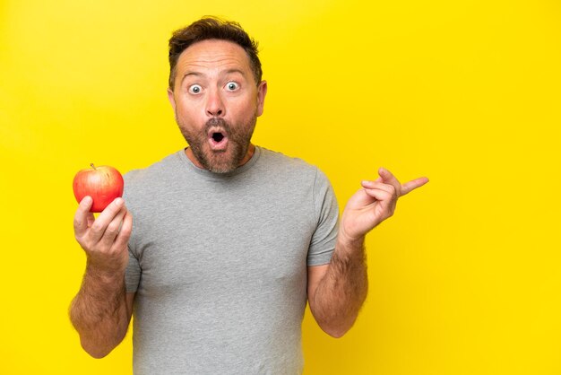 Foto blanke man van middelbare leeftijd met een appel geïsoleerd op gele achtergrond verrast en wijzende kant