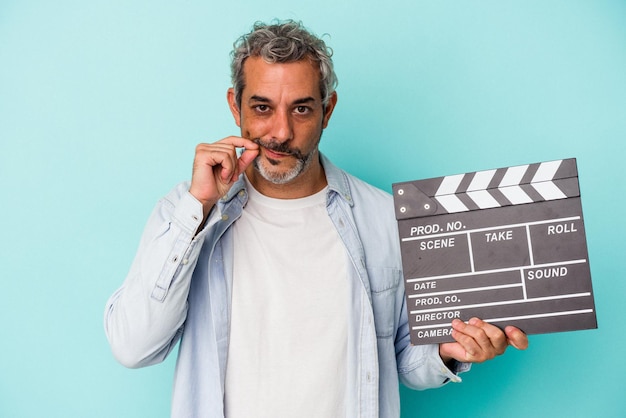 Blanke man van middelbare leeftijd met een Filmklapper geïsoleerd op een blauwe achtergrond met vingers op de lippen die een geheim houden.