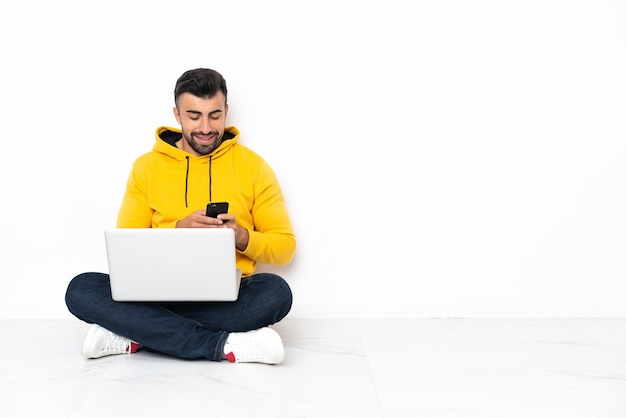 Blanke man zittend op de vloer met zijn laptop een bericht te sturen met de gsm