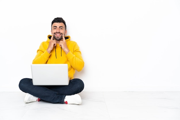 Blanke man zittend op de vloer met zijn laptop lachend met een gelukkige en aangename uitdrukking