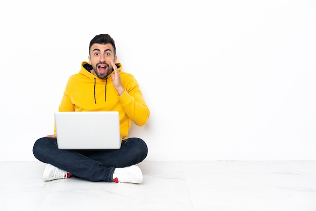 Blanke man zittend op de vloer met zijn laptop met verbazing en geschokt gelaatsuitdrukking