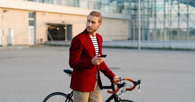 Blanke mannelijke fietser in casual stijl en rode jas toonaangevende fiets en tikken of scrollen op smartphone op straat. Knappe jongeman wandelen met fiets en SMS-bericht op mobiele telefoon.