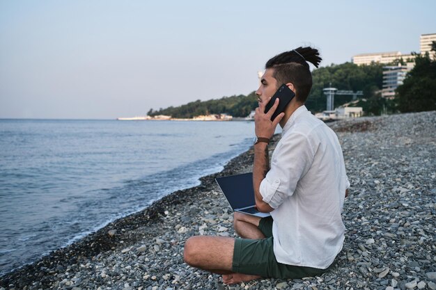 Blanke mannelijke freelancer zit op het strand en werkt op afstand online met een laptop