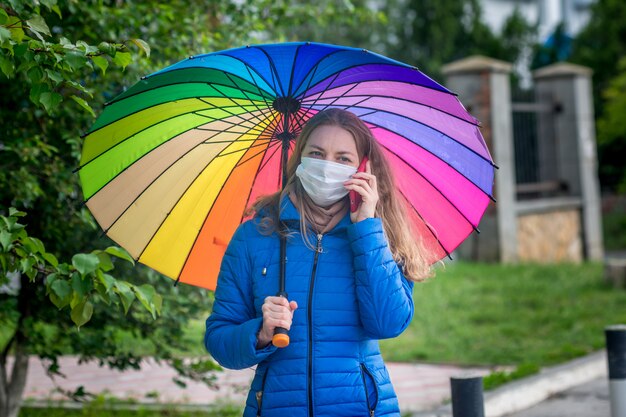 Blanke meisje in een beschermend masker staat op een lege straat bij een bushalte onder een paraplu in de lente regen en praat over de telefoon.