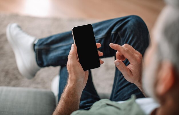 Blanke oude man met baard kijkt naar videospel op smartphone met leeg scherm in woonkamer