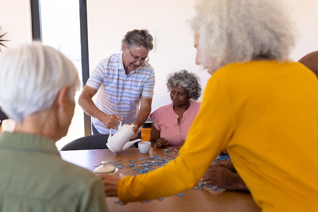 Blanke senior man die koffie serveert aan multiraciale vrienden die puzzel spelen in een verpleeghuis. Spel, verwarring, brainstormen, onveranderd, samenzijn, ondersteuning, begeleid wonen en pensioen.