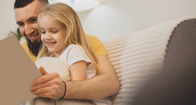 Blanke vader met grote baard die zijn meisje leert lezen met behulp van een boek dat op de bank ligt