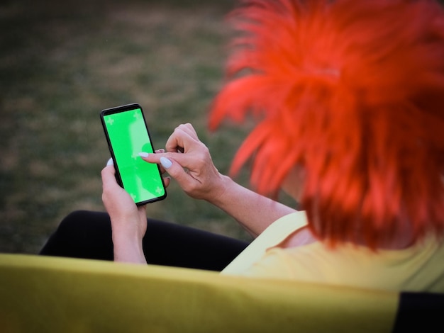 Blanke vrouw die een pruik met Belgische vlag draagt en een smartphone vasthoudt met een groen scherm dat er met haar vinger naar wijst